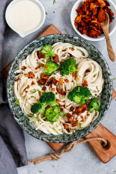 Vegane Spaghetti Carbonara angerichtet mit Kokos-Speck und etwas Brokkoli in einer Marmorfarbenen Schüssel auf Holzbrett