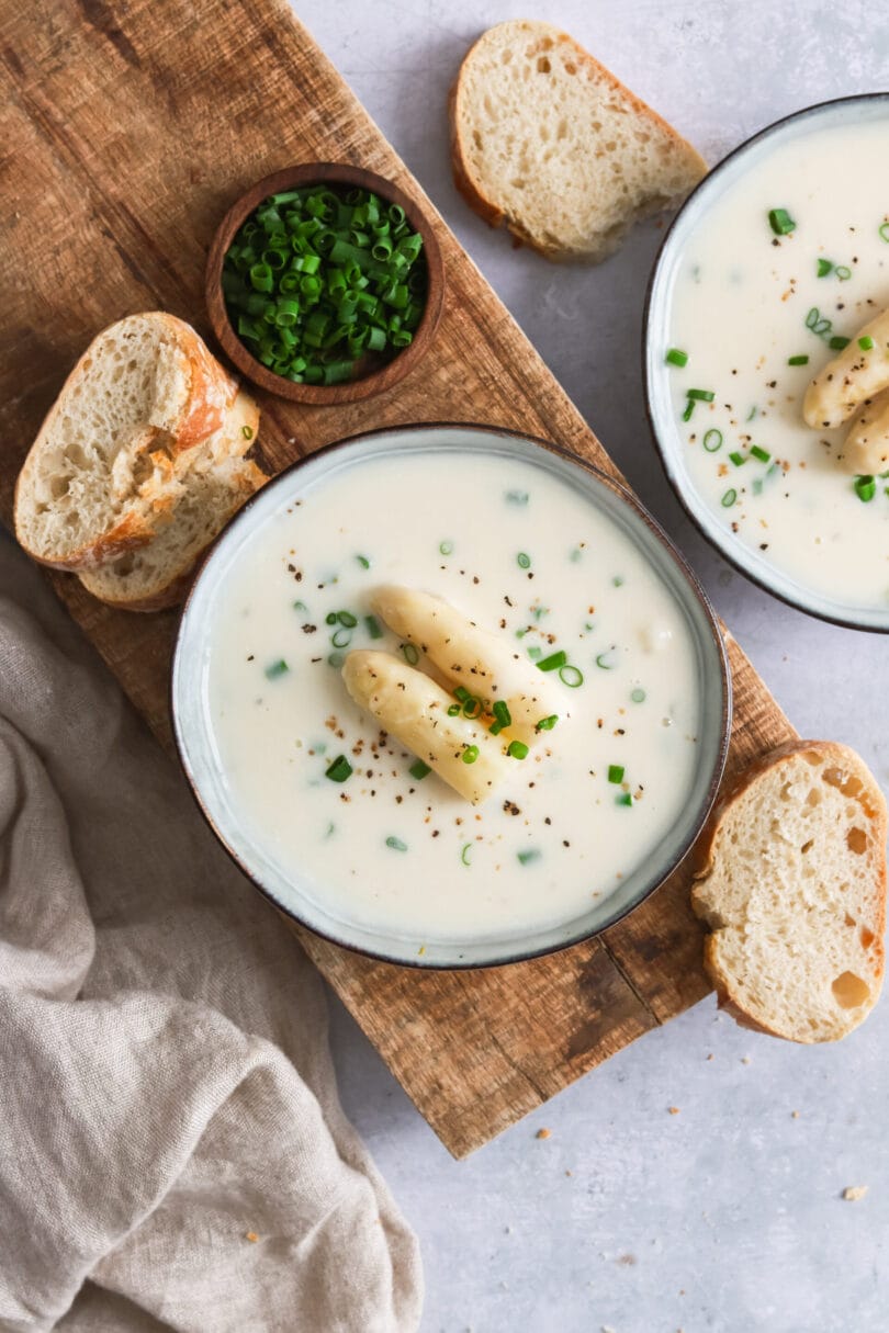 Zwei Suppenschüsseln mit veganer Spargelcremesuppe & Brot auf einem Schneidebrett