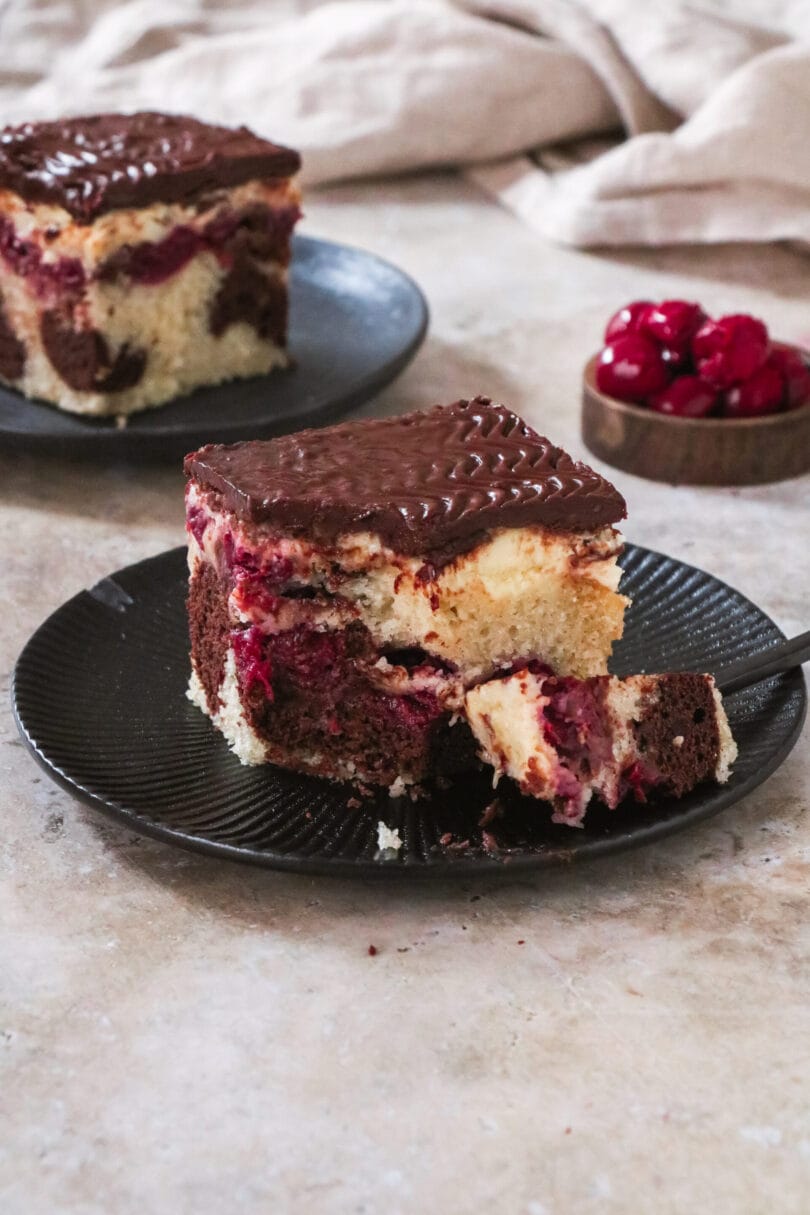 Ein Stück veganer Donauwellen-Kuchen, bestehend aus hellem und dunklem Teig mit Kirschen und Schokoladenglasur.