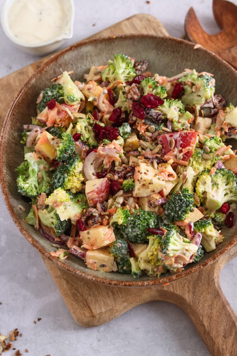 Gesunder Brokkoli-Apfel-Salat mit Cranberries, Karotten, Zwiebeln und Sonnenblumenkernen in einer cremigen Joghurtdressing-Schüssel, serviert auf einem Holzbrett.