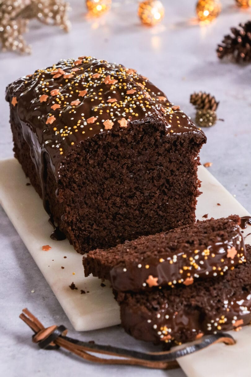 Veganer Gewürzkuchen mit Schokoladenglasur und Sternenstreuseln auf einer weißen Platte, daneben zwei angeschnittene Stücke, weihnachtliche Deko im Hintergrund.