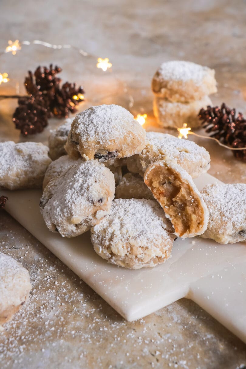 Veganes Stollenkonfekt mit Puderzucker bestäubt, arrangiert auf einer weißen Platte mit weihnachtlicher Beleuchtung und Deko aus Tannenzapfen im Hintergrund.