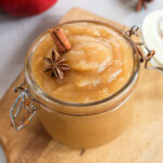 Nahaufnahme eines Glases mit cremigem Apfelmus, dekoriert mit Zimtstange und Sternanis, auf einem Holzbrett.