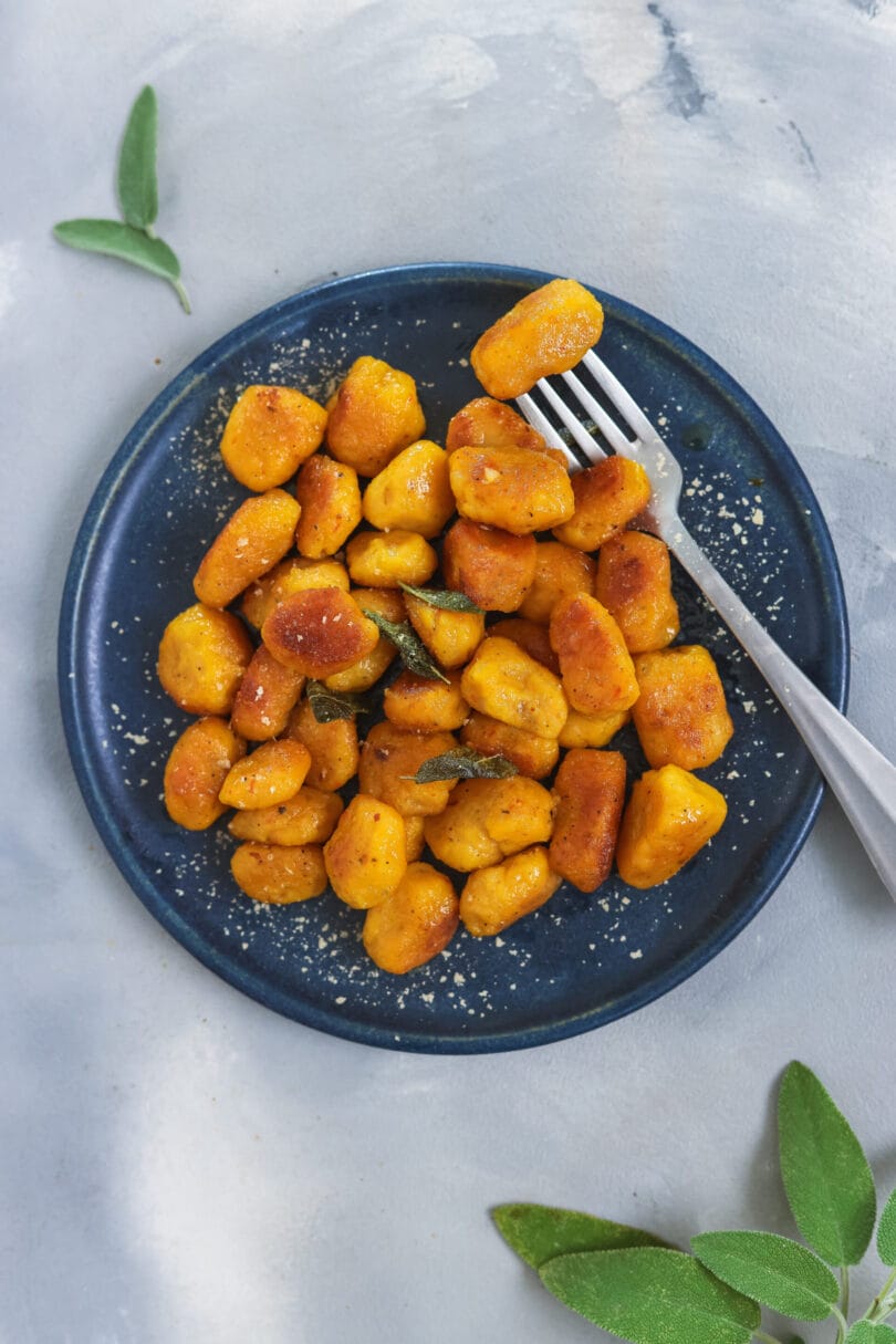 Hausgemachte, vegane Kürbis Gnocchi auf dunkelblauem Teller, knusprig angebraten mit Salbeiblättern und Parmesan, herbstliches Gericht.
