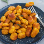 Hausgemachte, vegane Kürbis Gnocchi auf dunkelblauem Teller, knusprig angebraten mit Salbeiblättern und Parmesan, herbstliches Gericht.