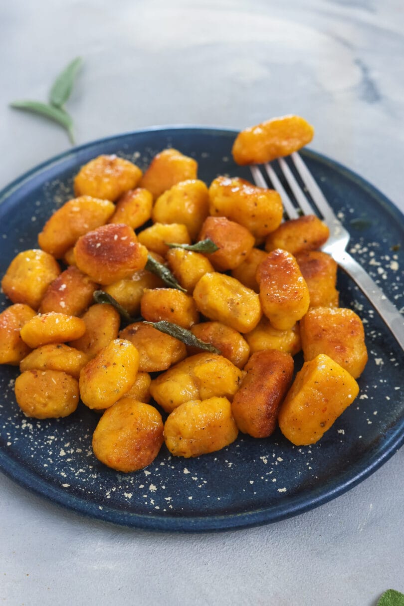 Hausgemachte, vegane Kürbis Gnocchi auf dunkelblauem Teller, knusprig angebraten mit Salbeiblättern und Parmesan, herbstliches Gericht.