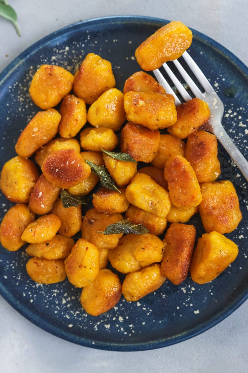 Hausgemachte, vegane Kürbis Gnocchi auf dunkelblauem Teller, knusprig angebraten mit Salbeiblättern und Parmesan, herbstliches Gericht.