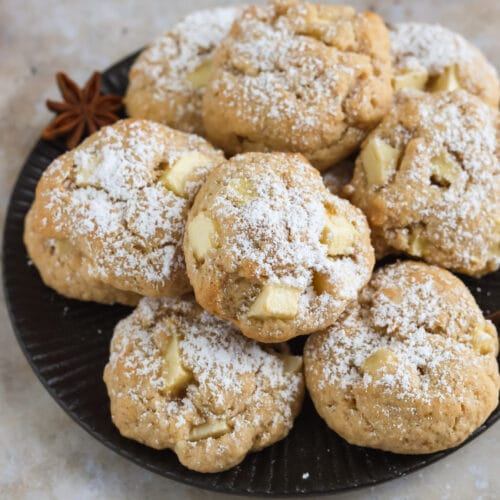 Saftige, vegane Apfelkekse mit Zimt auf einem dunklen Teller mit Puderzucker bestreut und Zimtsternen im Hintergrund
