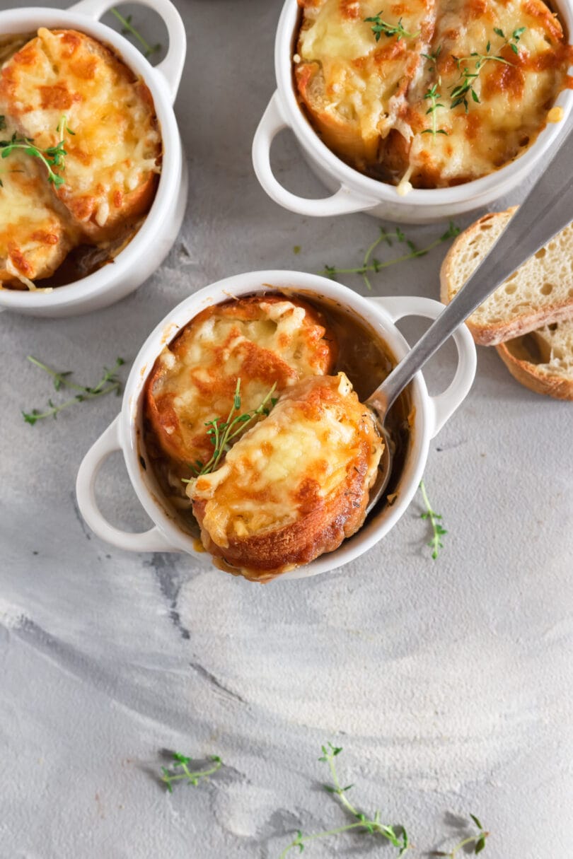 Mehrere weiße Suppenschalen mit französischer Zwiebelsuppe, goldbraun mit geschmolzenem Käse überbacken und mit Thymian garniert; daneben liegen Scheiben von Baguette.