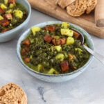 Veganer Grünkohleintopf aus gehacktem Grünkohl, Kartoffelwürfeln und angebratenem Tofu in saftiger Brühe, serviert auf hellem Untergrund mit rustikalem Brot daneben.