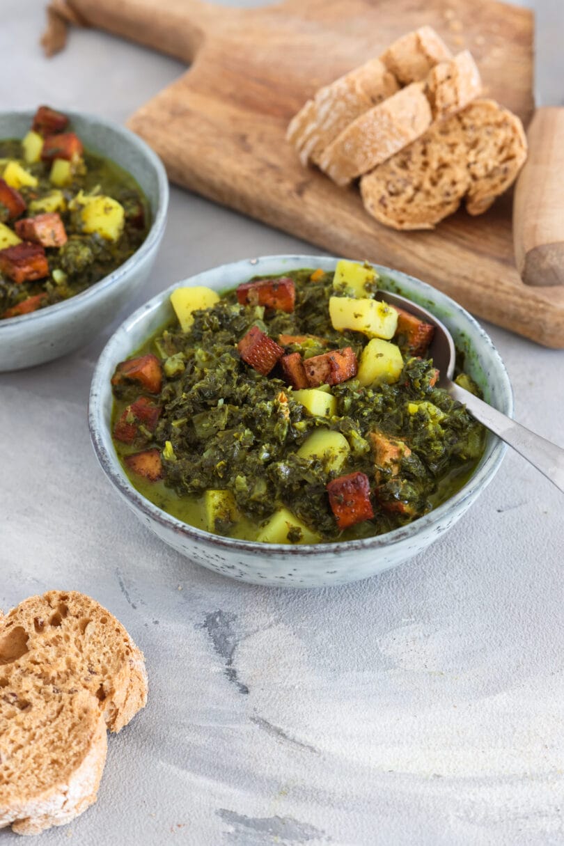 Veganer Grünkohleintopf aus gehacktem Grünkohl, Kartoffelwürfeln und angebratenem Tofu in saftiger Brühe, serviert auf hellem Untergrund mit rustikalem Brot daneben.