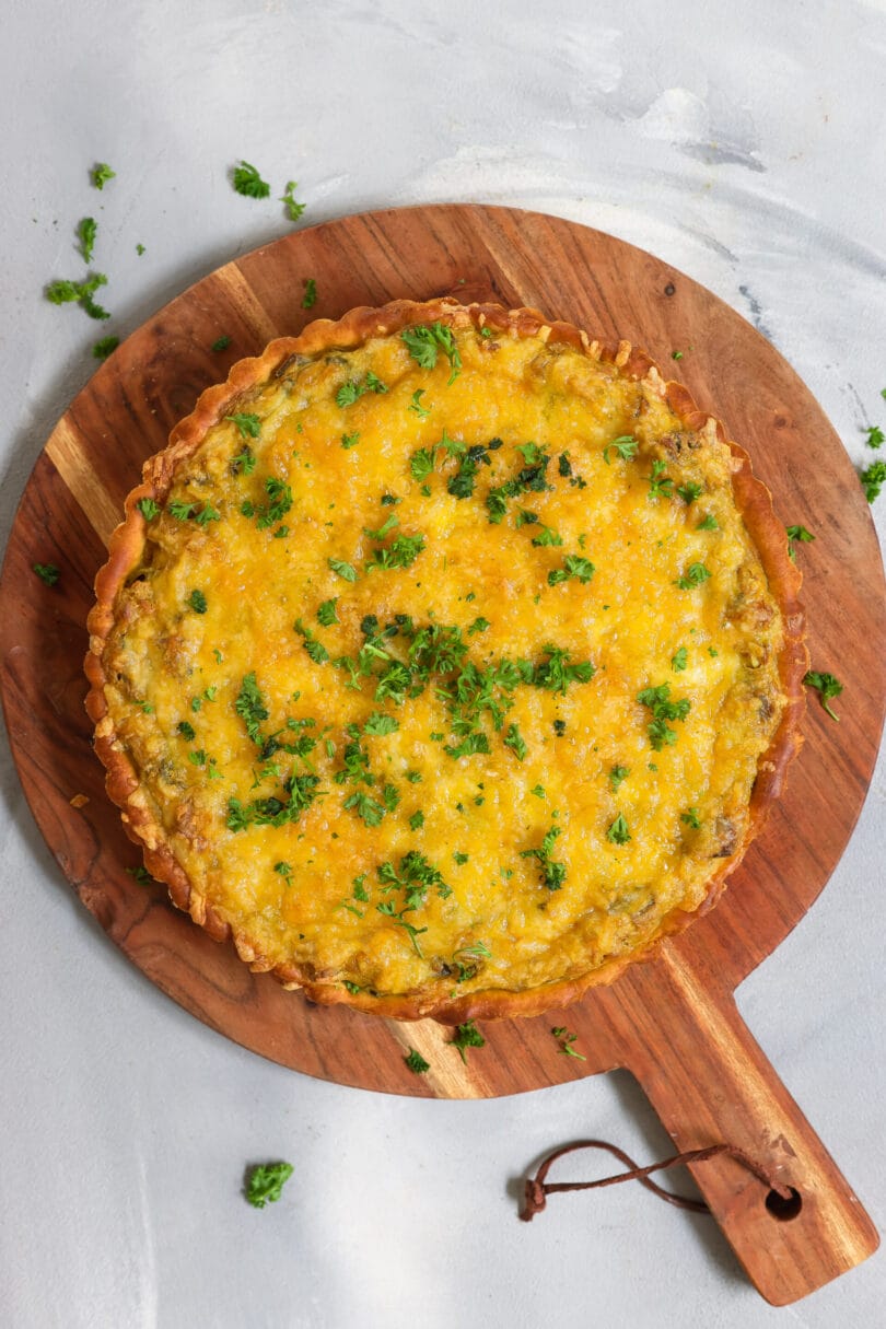 Ein goldbraun gebackene, herzhafte Quiche mit knusprigem Rand, belegt mit veganem Käse und frischer Petersilie, serviert auf einem runden Holzbrett vor hellem Hintergrund.