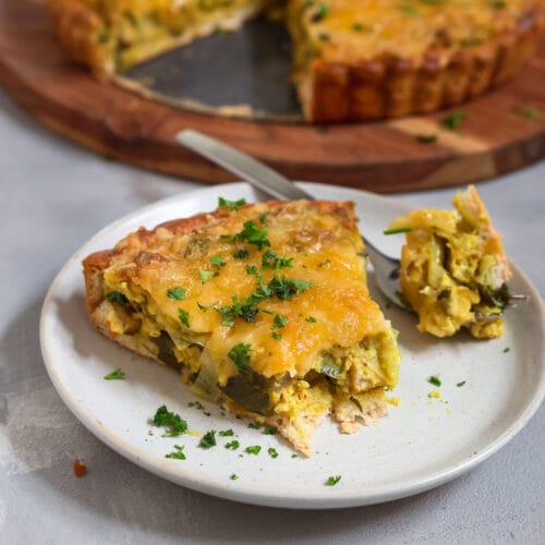 Ein goldbraun gebackene, herzhafte Quiche mit knusprigem Rand, belegt mit veganem Käse und frischer Petersilie, serviert auf einem runden Holzbrett vor hellem Hintergrund.