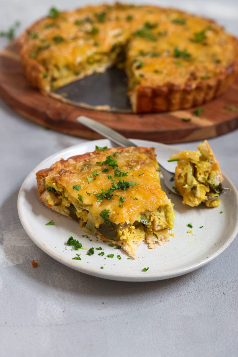 Ein goldbraun gebackene, herzhafte Quiche mit knusprigem Rand, belegt mit veganem Käse und frischer Petersilie, serviert auf einem runden Holzbrett vor hellem Hintergrund.