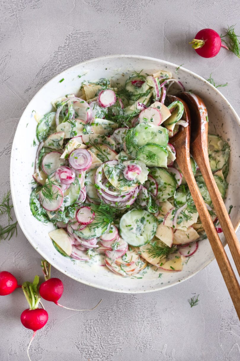 Veganer Radieschensalat mit Apfel, Gurke und cremigem Joghurtdressing in einer weißen Schüssel mit Holzlöffeln.