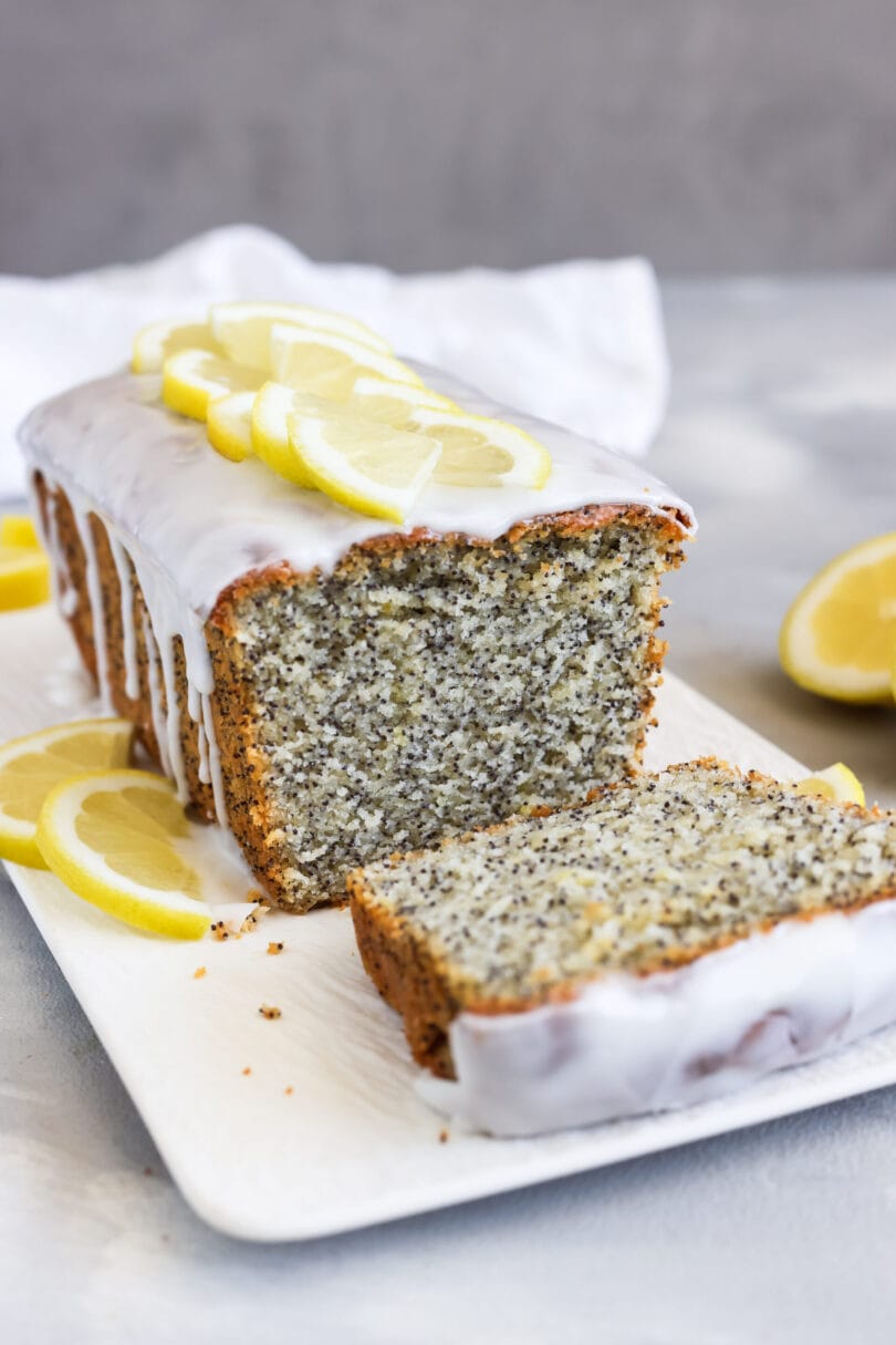 Saftiger Zitronen-Mohn-Kuchen mit Zitronenglasur und Zitronenscheiben, aufgeschnitten auf einem weißen Servierbrett.