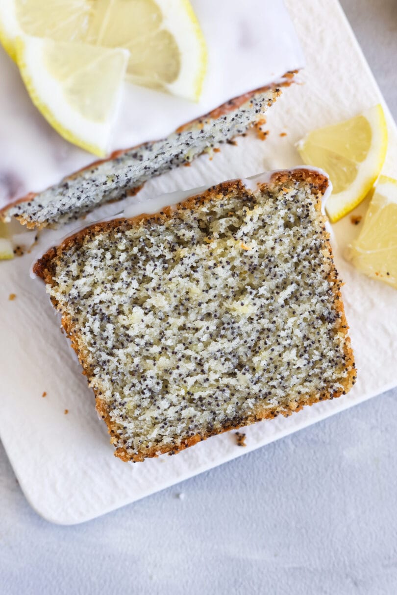 Saftiger veganer Zitronen-Mohn-Kuchen mit Puderzuckerglasur, angeschnitten auf einem hellen Servierbrett, dekoriert mit frischen Zitronenscheiben.