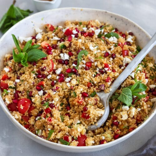 Veganer Couscous-Salat mit Kichererbsen, Cherrytomaten, Gurke, Granatapfelkernen, veganem Feta und frischer Minze in einer großen Schüssel mit Servierlöffel.