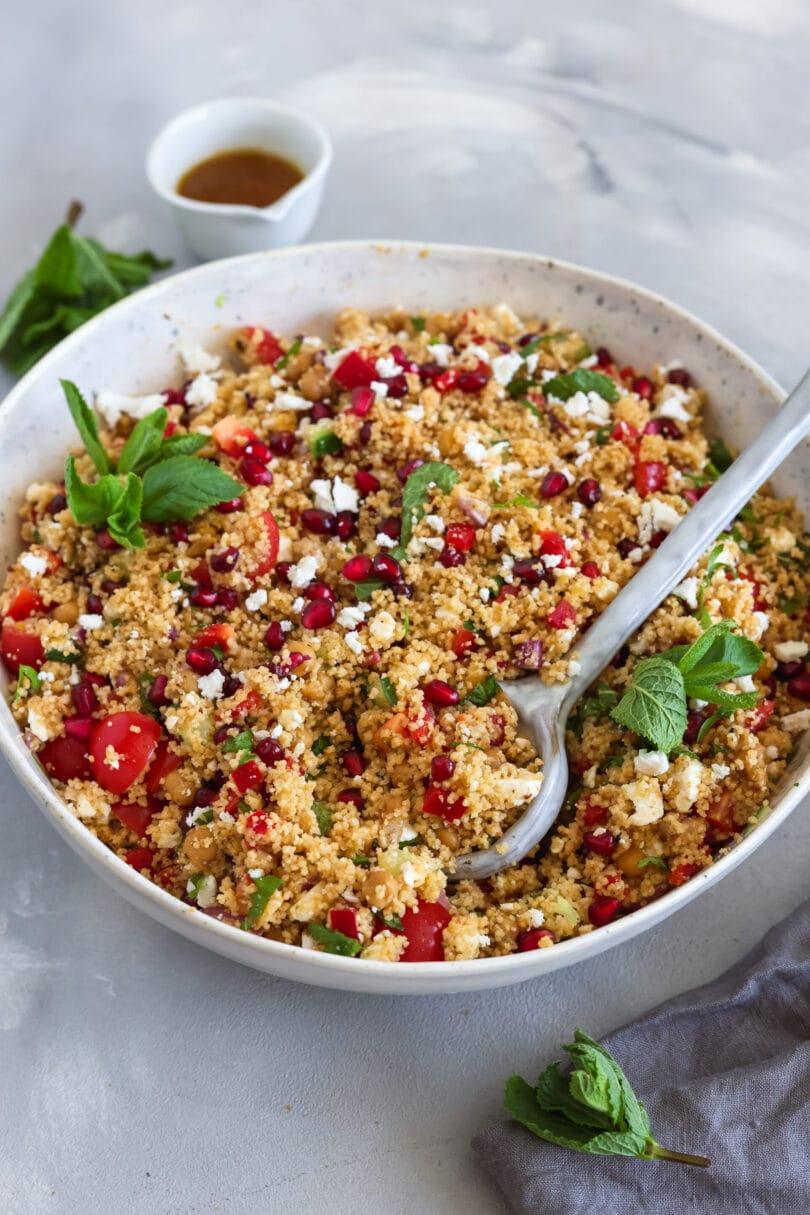Veganer Couscous-Salat mit Kichererbsen, Cherrytomaten, Gurke, Granatapfelkernen, veganem Feta und frischer Minze in einer großen Schüssel mit Servierlöffel.