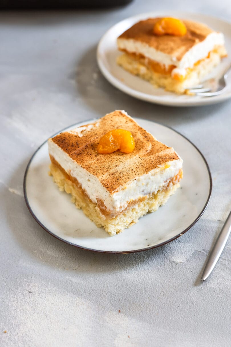 Ein Stück veganer Fantakuchen mit saftigem Rührteig, Mandarinen und cremiger Sahneschicht, bestäubt mit Zimt und garniert mit einer Mandarine auf einem Dessertteller.