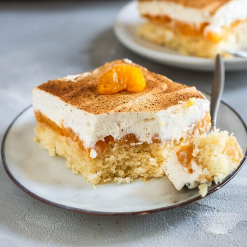 Nahaufnahme eines veganen Fantakuchens mit fluffigem Boden, Mandarinen und cremigem Topping, mit Zimt bestäubt und einer Mandarine als Dekoration.