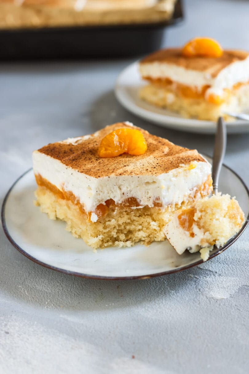 Nahaufnahme eines veganen Fantakuchens mit fluffigem Boden, Mandarinen und cremigem Topping, mit Zimt bestäubt und einer Mandarine als Dekoration.