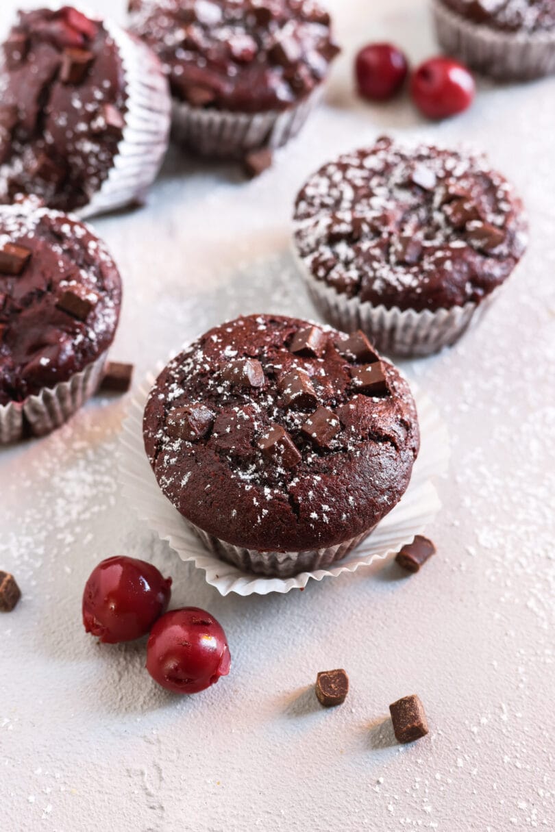 Saftiger veganer Schoko-Kirsch-Muffin mit Schokostückchen und Puderzucker auf hellem Untergrund mit Kirschen.