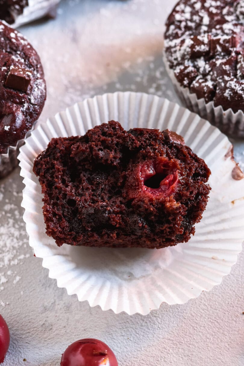 Veganer Schoko-Kirsch-Muffin mit saftiger Krume und ganzer Kirsche im Inneren, halbiert in einer Papierförmchen.