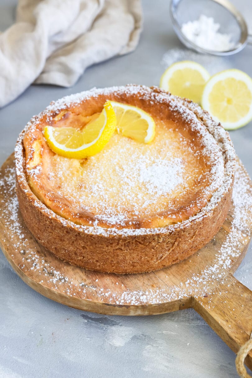 Ganzer veganer Zitronen-Käsekuchen mit goldbrauner Oberfläche, knusprigem Boden, Puderzucker und frischen Zitronenscheiben auf einem Holzbrett.