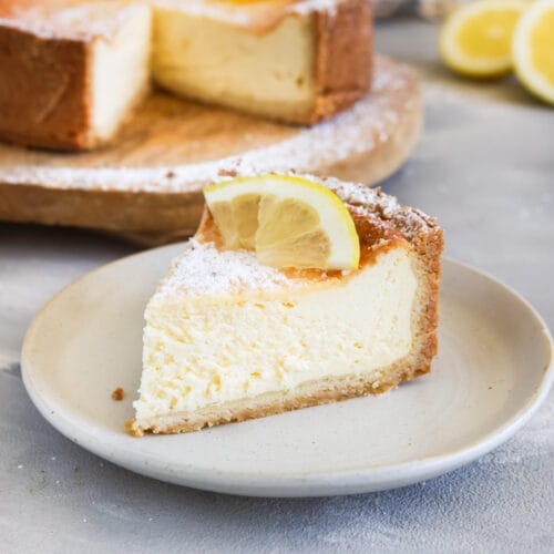 Cremiger veganer Zitronen-Käsekuchen mit knusprigem Boden und frischer Zitronengarnitur, serviert als Kuchenstück vor dem angeschnittenen Kuchen.