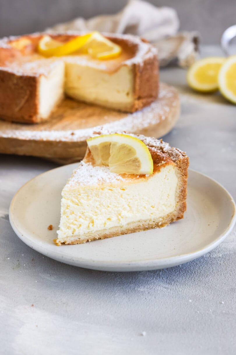 Cremiger veganer Zitronen-Käsekuchen mit knusprigem Boden und frischer Zitronengarnitur, serviert als Kuchenstück vor dem angeschnittenen Kuchen.