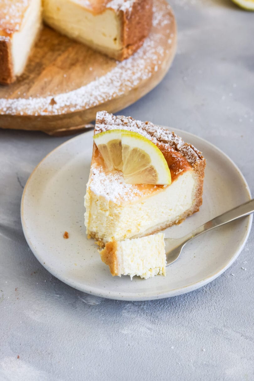 Ein Stück veganer Zitronen-Käsekuchen mit cremiger Füllung, knusprigem Boden, Puderzucker und Zitronenscheibe auf einem weißen Teller.