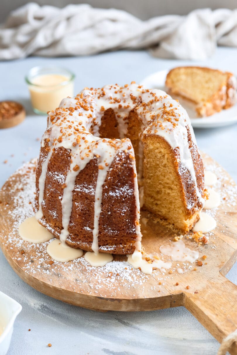 Angeschnittener veganer Eierlikörkuchen als Gugelhupf mit weißem Guss, Krokant und Puderzucker auf einem runden Holzbrett.