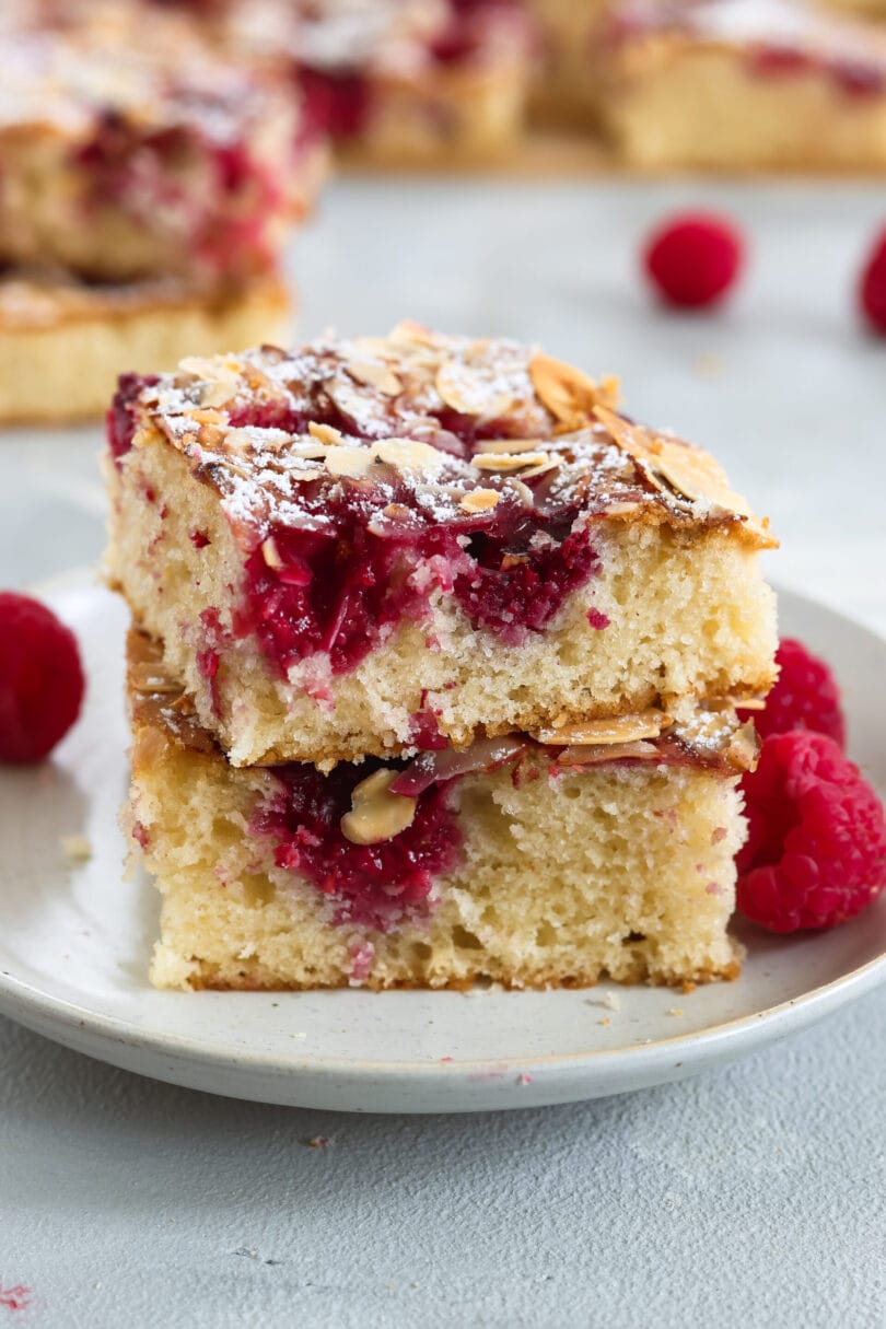 Zwei Stücke veganer Himbeer-Blechkuchen mit frischen Himbeeren, knusprigen Mandelblättchen und Puderzucker auf einem Teller.