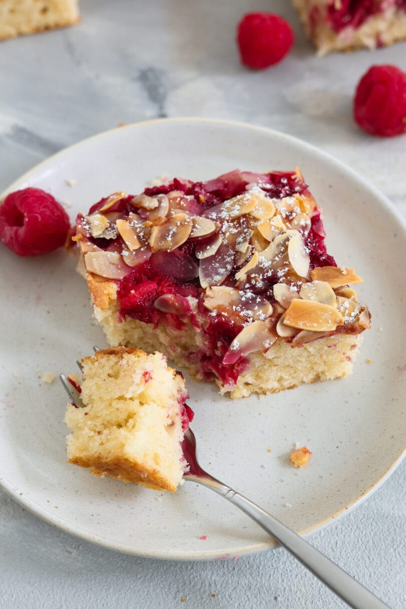 Saftiges Stück veganer Himbeer-Blechkuchen mit gerösteten Mandelblättchen und Puderzucker auf einem hellen Teller.