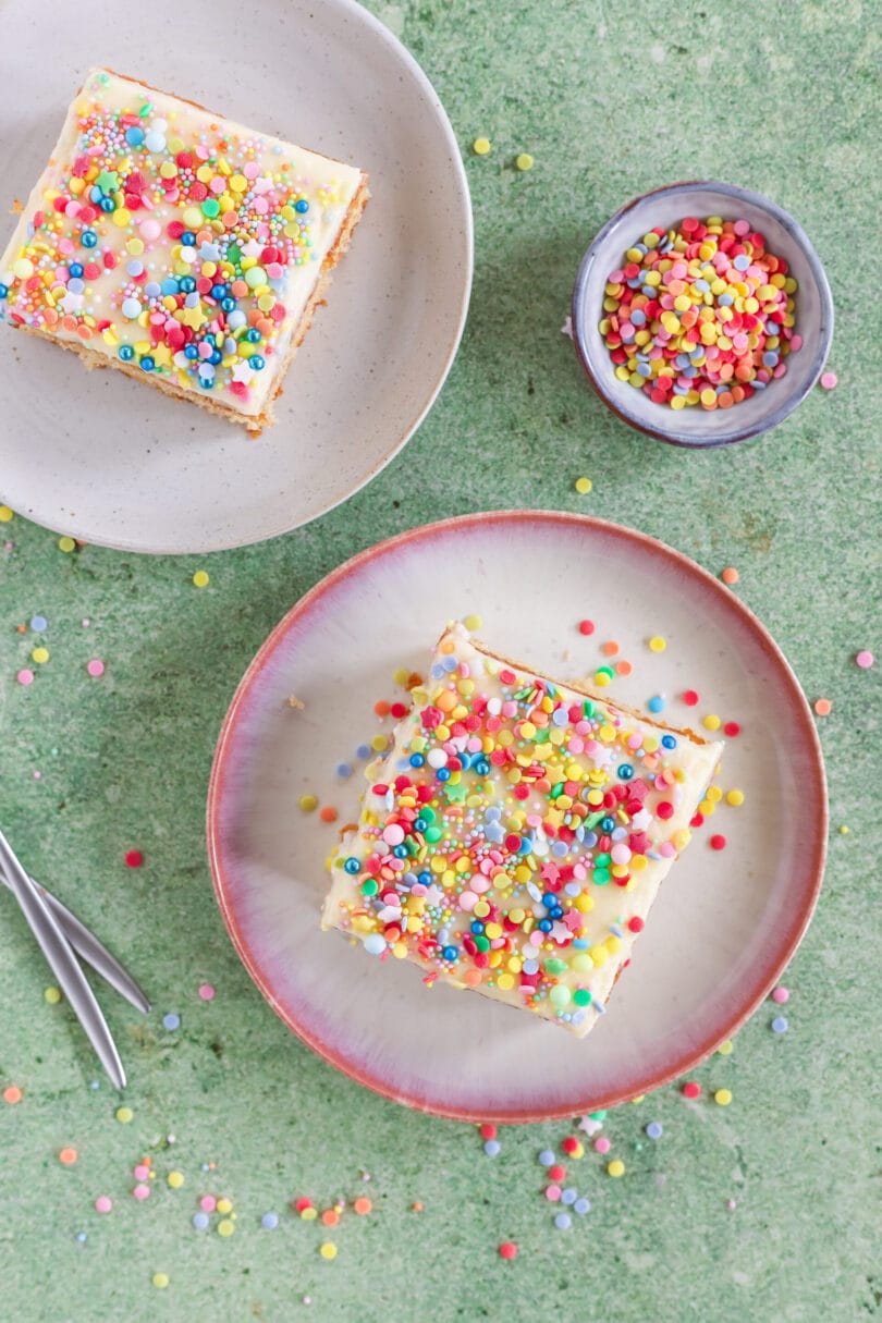 Veganer Konfettikuchen mit bunten Streuseln auf einem Teller, daneben ein weiteres Kuchenstück und eine Schale mit Zuckerstreuseln.