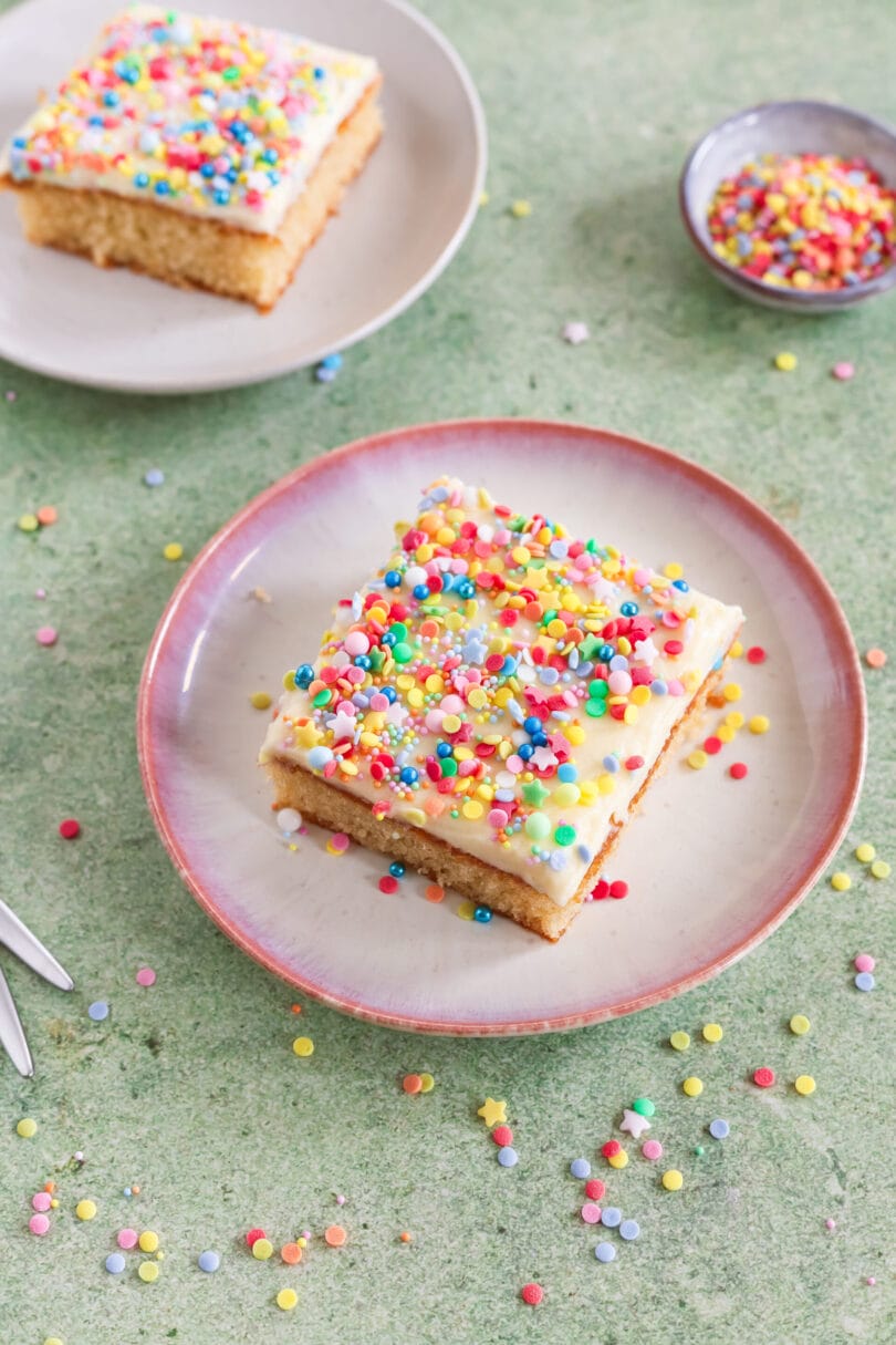 Veganer Konfettikuchen mit cremigem Frosting und bunten Zuckerstreuseln auf einem Teller vor grünem Hintergrund.
