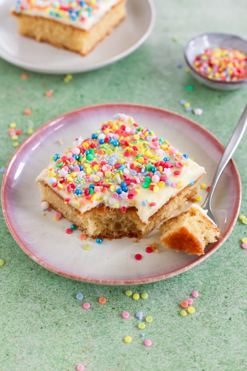 Stück veganer Konfettikuchen mit hellem Frosting und bunten Streuseln auf einem Teller, daneben eine Kuchengabel.