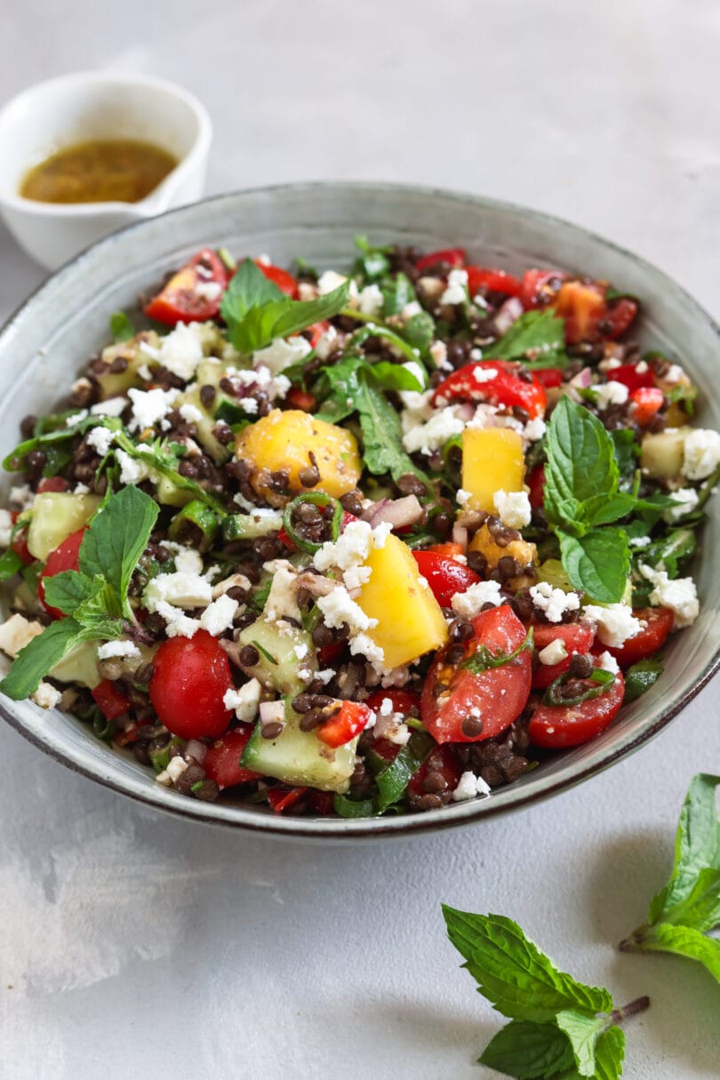 Nahaufnahme von veganem Belugalinsensalat mit Mango, veganem Feta, Kirschtomaten, Gurke und frischen Kräutern.