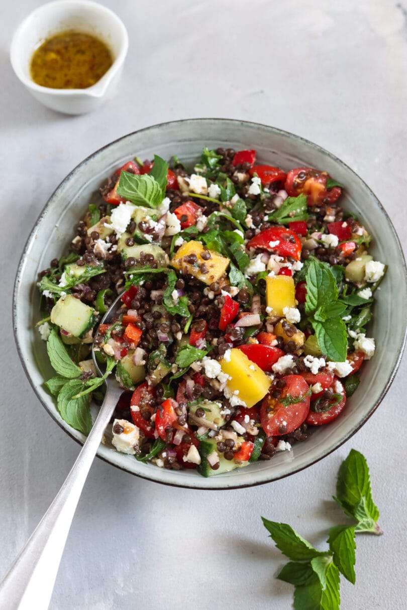 Bunter veganer Belugalinsensalat mit Mango und Feta, dazu Tomaten, Gurke, Paprika und frische Minze.