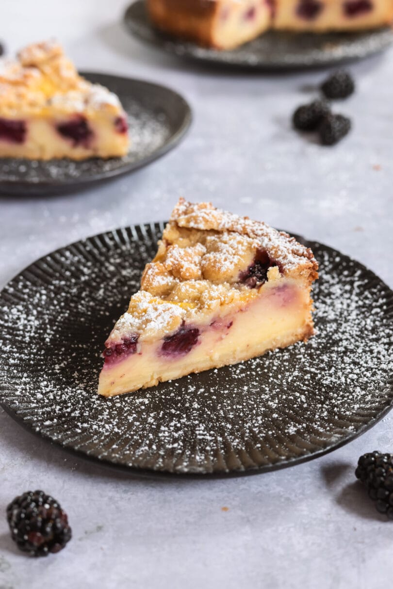 Stück veganer Brombeerkuchen mit cremigem Pudding und Streuseln auf einem schwarzen Teller.