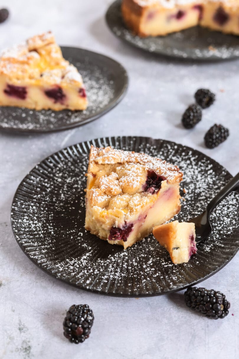 Veganer Brombeer-Pudding-Kuchen mit Streuseln und Puderzucker, serviert mit frischen Brombeeren.