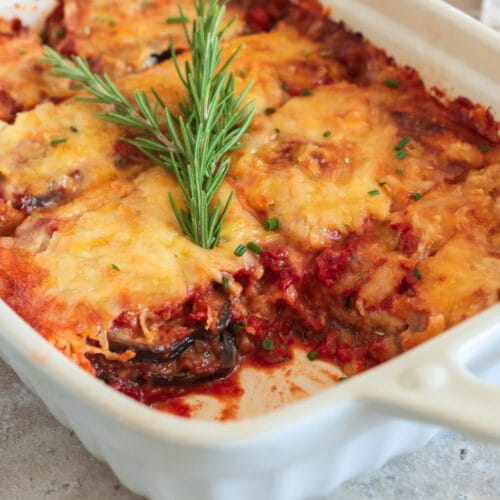 Vegane Parmigiana di Melanzane in weißer Auflaufform, überbacken mit goldbraunem veganem Käse und garniert mit Rosmarin.