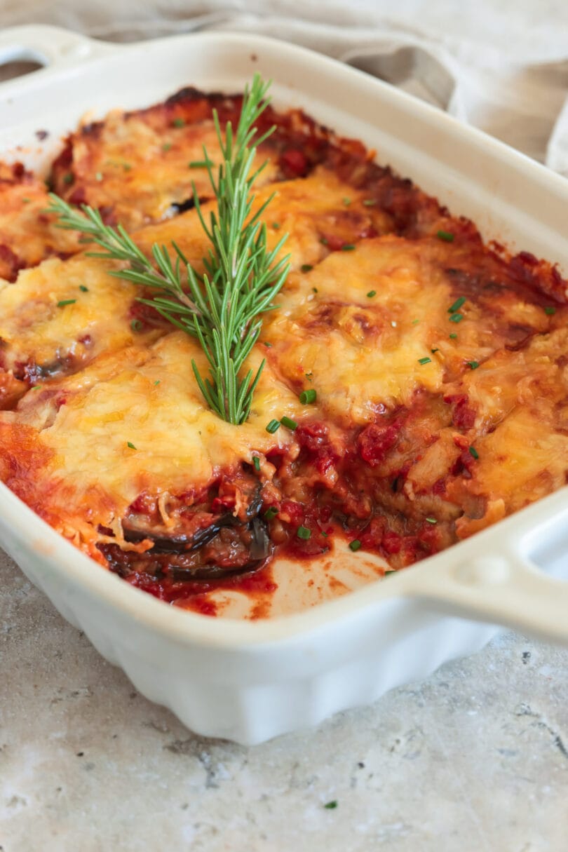 Vegane Parmigiana di Melanzane in weißer Auflaufform, überbacken mit goldbraunem veganem Käse und garniert mit Rosmarin.