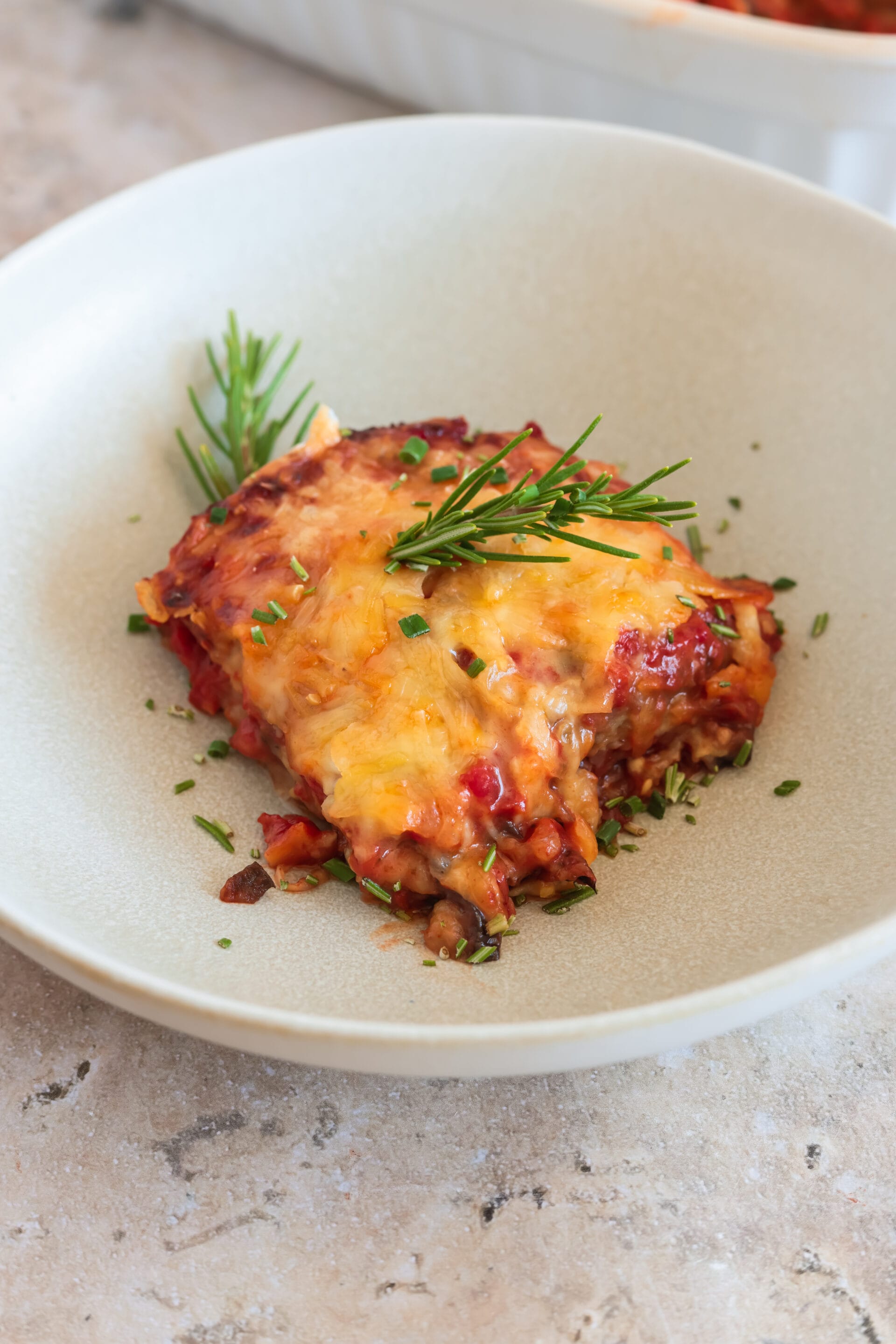 Eine Portion vegane Parmigiana di Melanzane auf einem hellen Teller, bestreut mit Schnittlauch und Rosmarin.