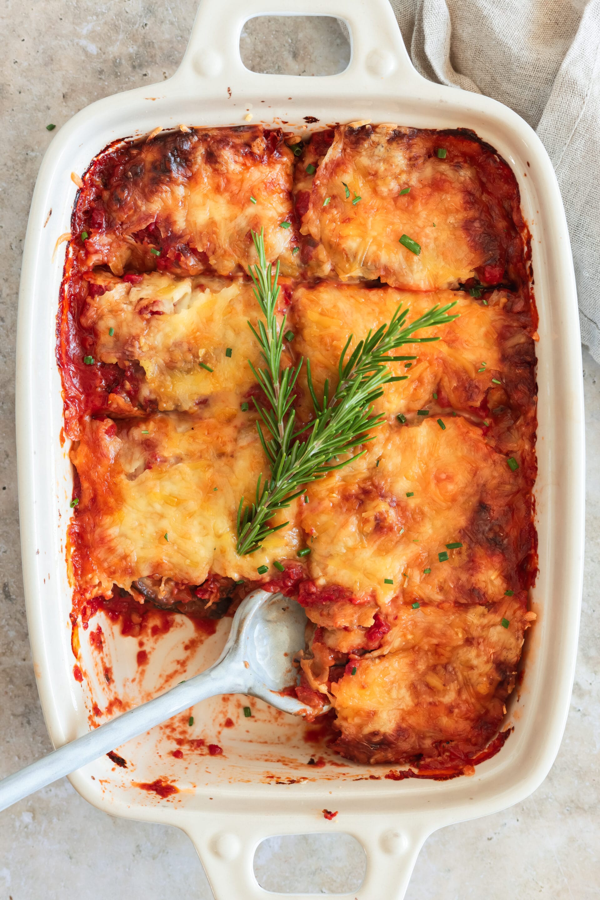 Auflaufform mit veganem Auberginenauflauf von oben, Rosmarinzweig auf dem goldbraunen Käse und Löffel zum Servieren.
