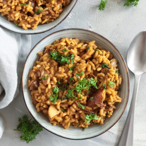 Zwei Schalen veganes Steinpilzrisotto mit Petersilie garniert, dazu Löffel und Leinentuch.