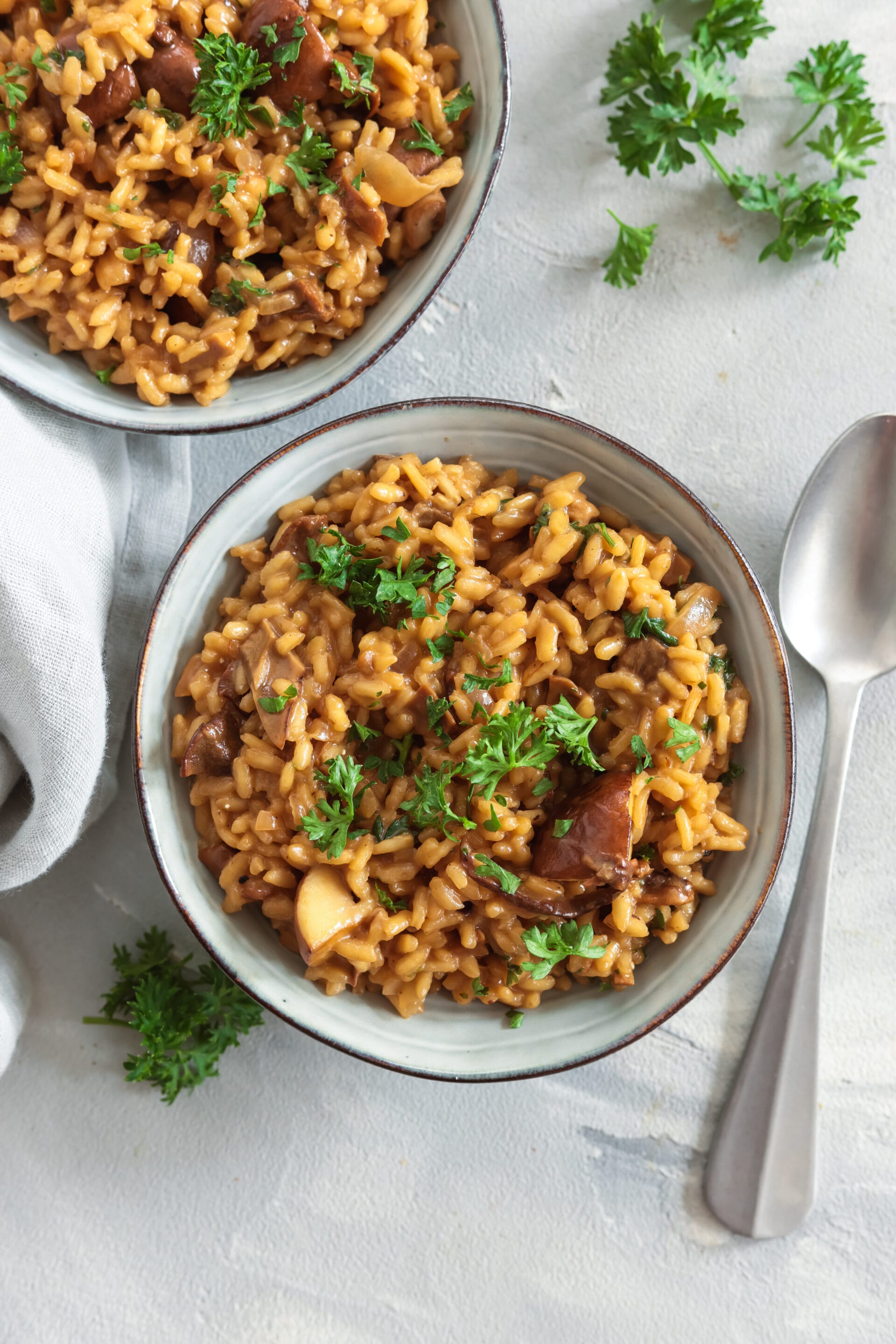 Zwei Schalen veganes Steinpilzrisotto mit Petersilie garniert, dazu Löffel und Leinentuch.